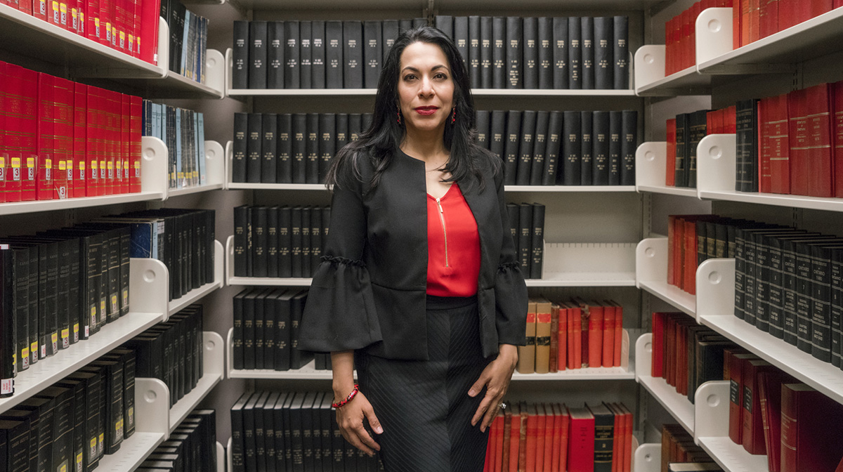 A woman, with long black hair, wearing a red shirt and black blazer and skirt, stands in a library aisle stacked with reference books.